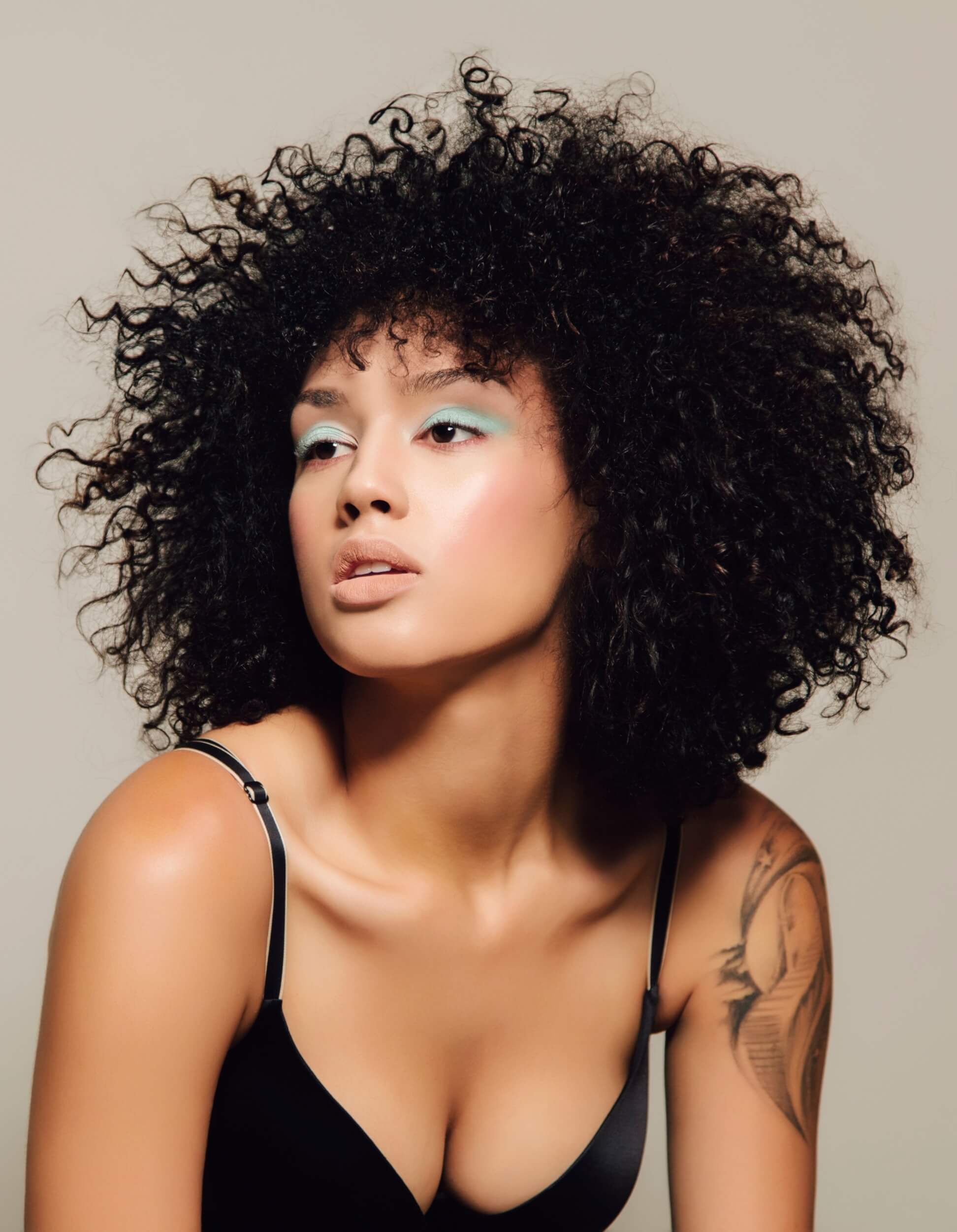 Feminist Threads fashion model with natural curly hair, bold turquoise eyeshadow and tattoo on arm wearing a black bra against a neutral background