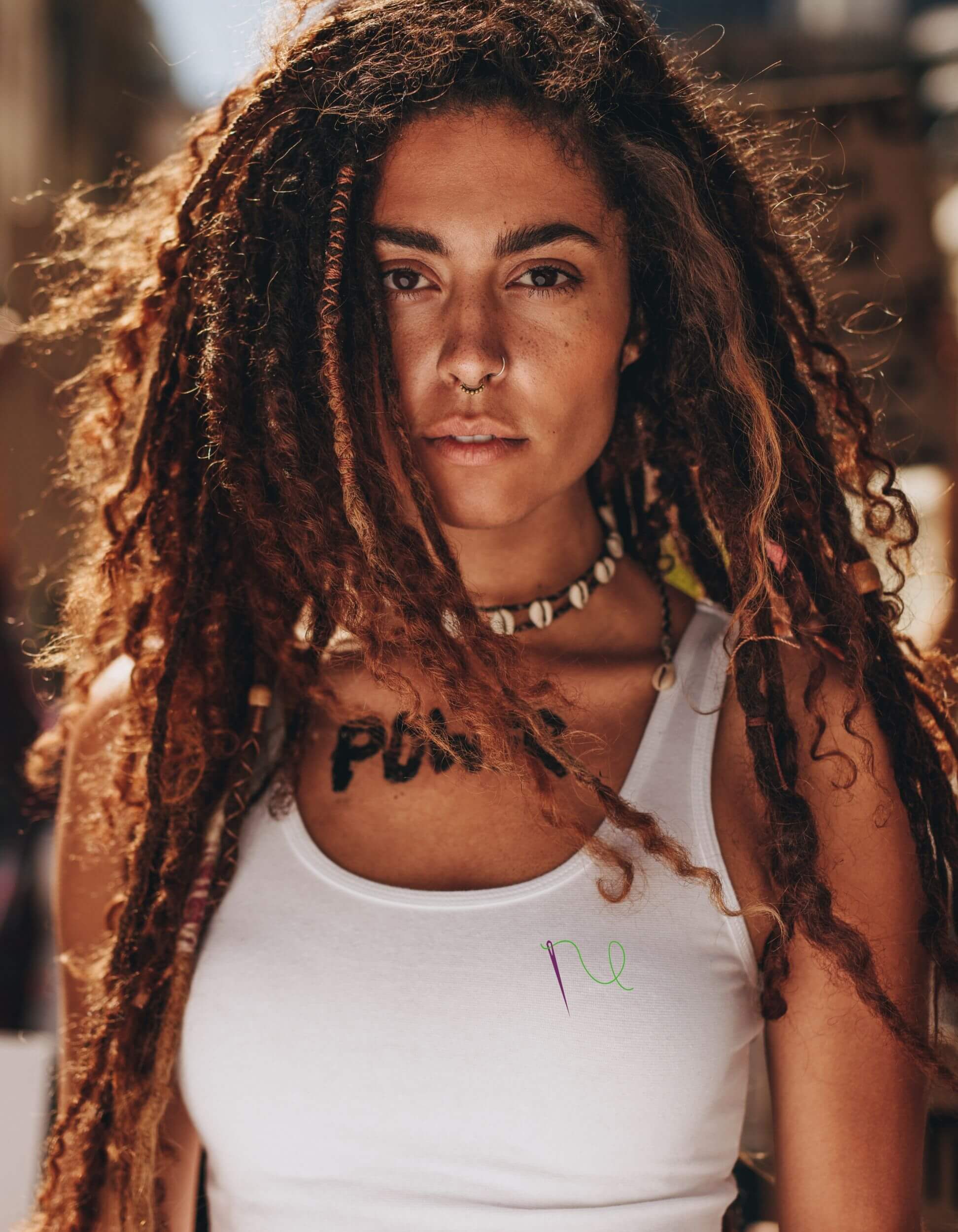 Woman with long dreadlocks, nose piercing, and 'power' written across her chest, wearing a white vest top with the Feminist Threads logo in suffragette colours at women’s march in London 
